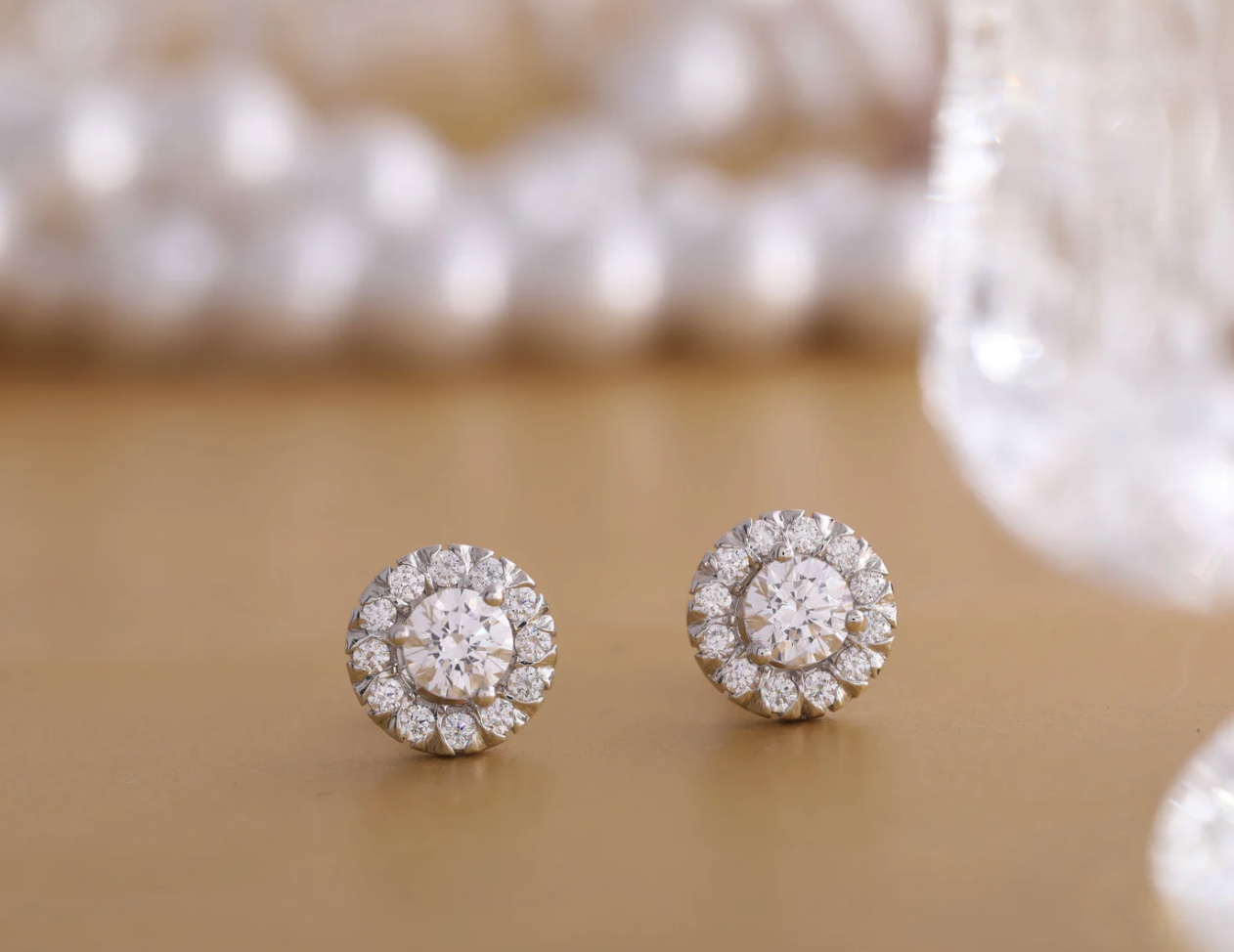 Pair of round diamond halo stud earrings on a warm neutral surface.